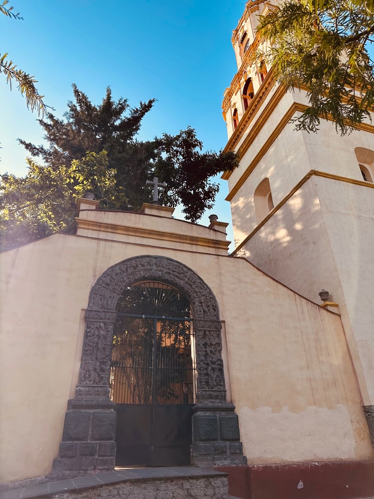 Historic colonial church exterior with ornate arched stone doorway, cream and terracotta walls, bell tower, and green foliage against blue sky