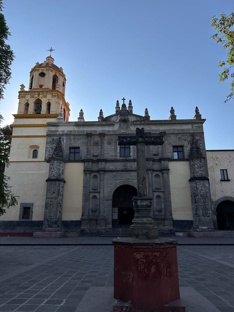 Historic colonial church with cream and stone facade, ornate bell tower, and decorative crest on roof against clear blue sky